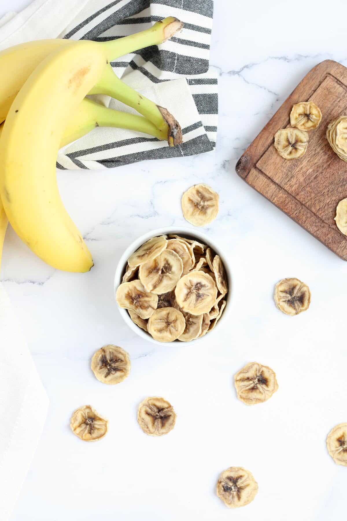 Top view of homemade banana chip on a marble surface, fresh bananas and wooden cutting board in the background
