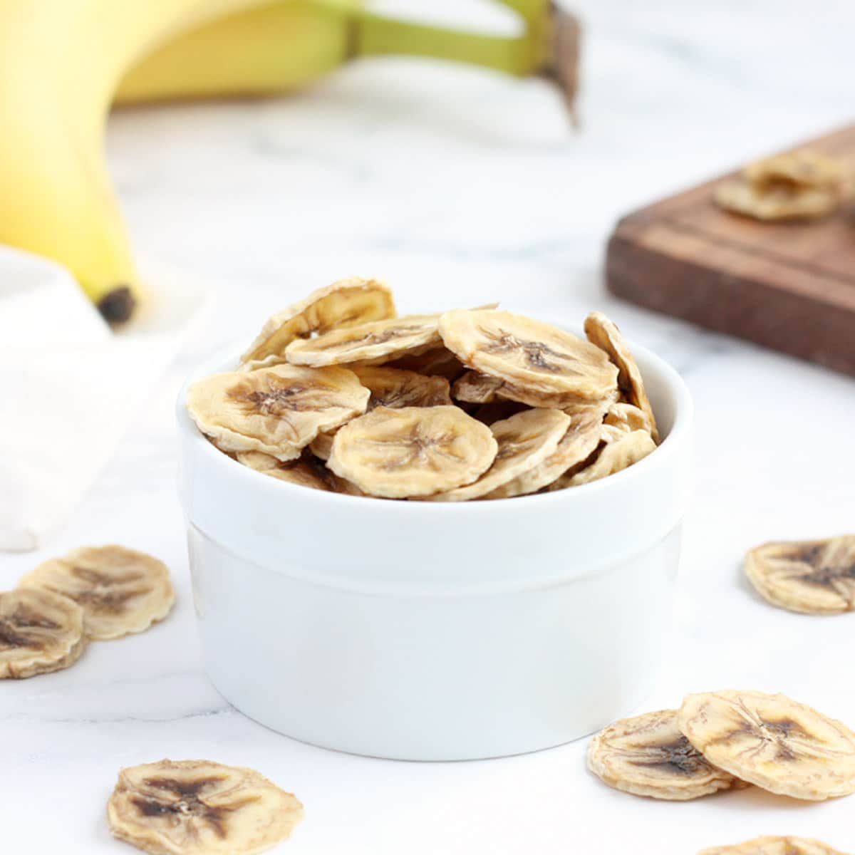 homemade banana chips in a white bowl with fresh bananas in the background