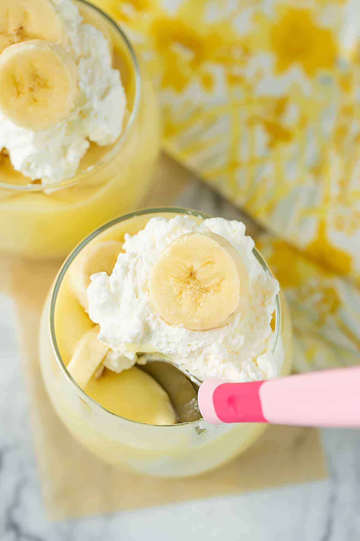 banana pudding recipe with whip cream, fresh bananas and a pink spoon in a glass filled with pudding