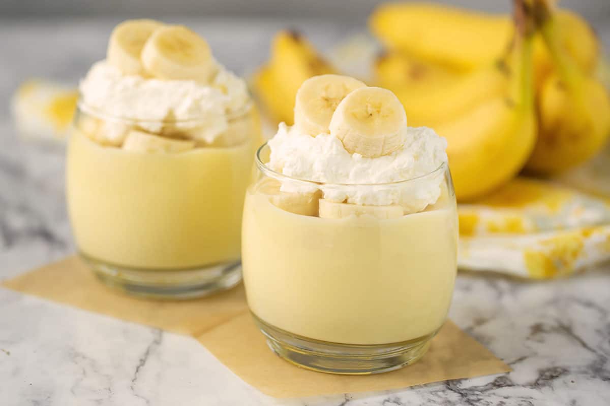 banana pudding in glasses with fresh bananas, whip cream and bananas in the background