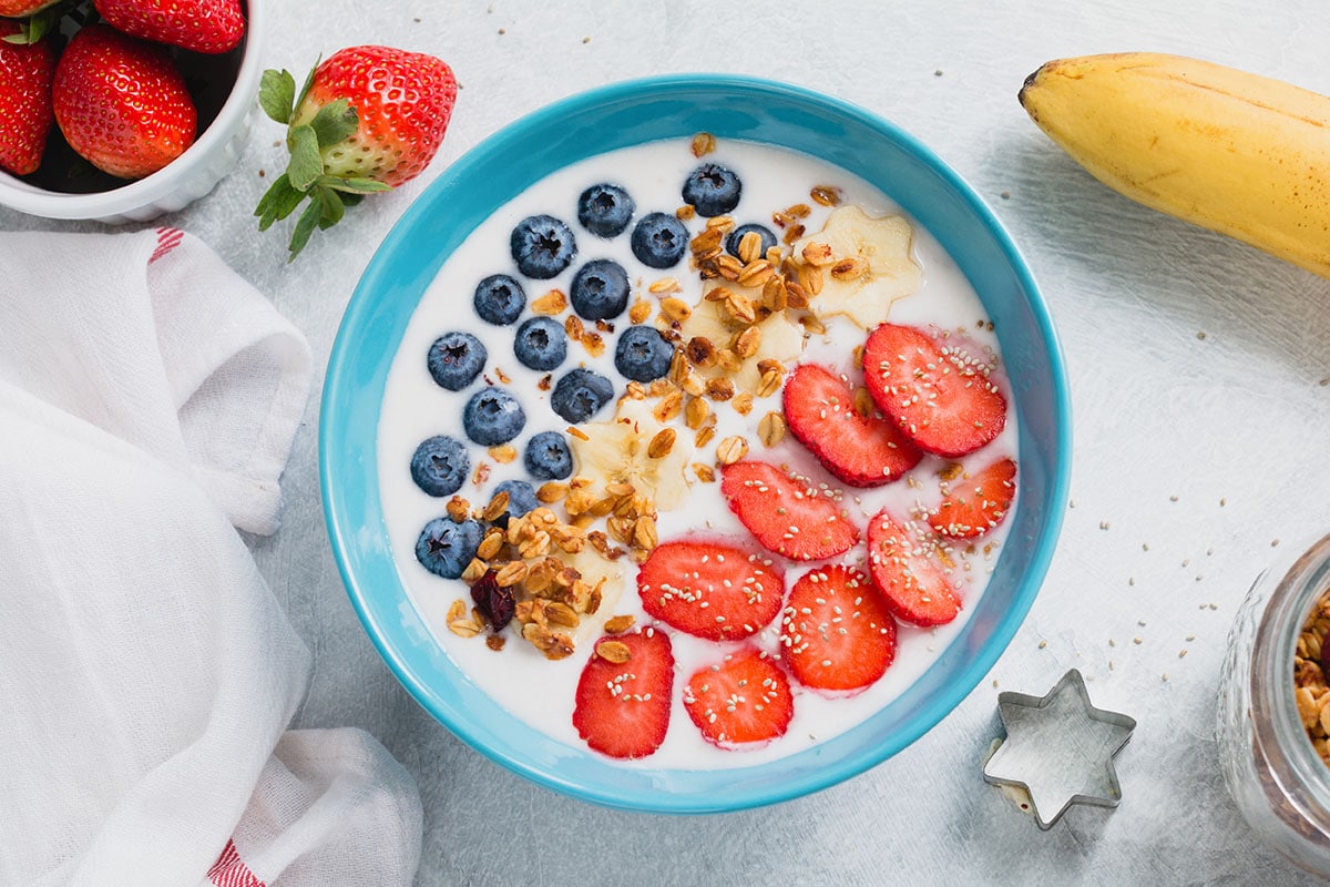 fruit and yogurt bowl with blueberries, strawberries, banana stars and granola in a blue bowl with strawberries, bananas, and a metal star cutter in the background