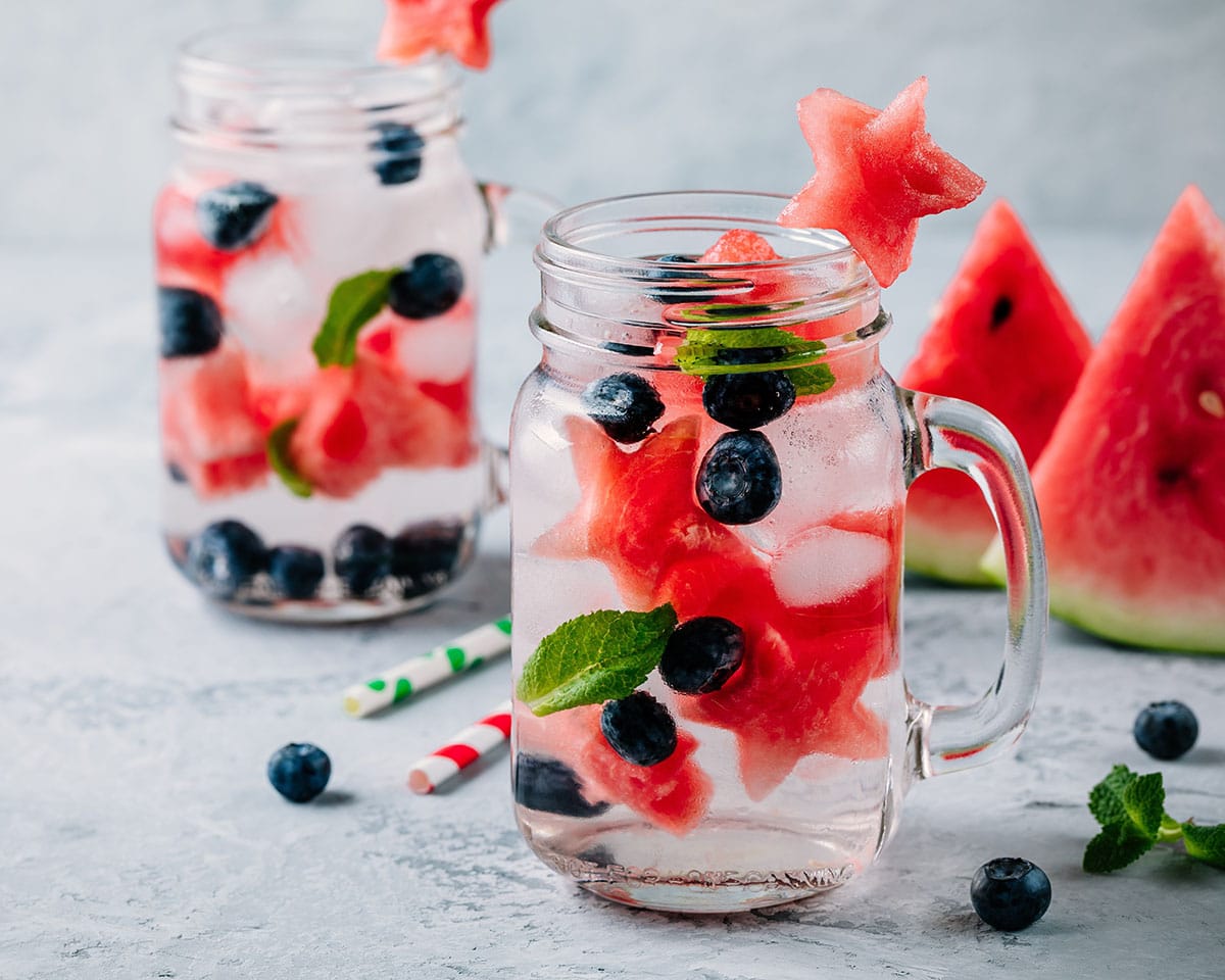 fruit infused water with blueberries, mint, watermelon stars, and ice in a glass mason jar with watermelon slices in the background.