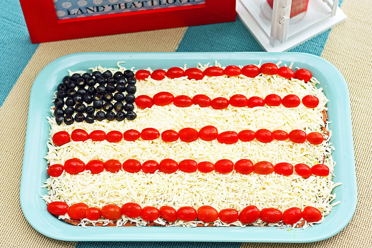 layered bean dip that looks like an american flag with tomatoes, mozzarella cheese and olives