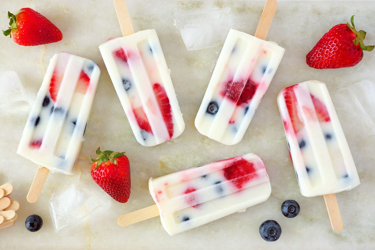 Healthy strawberry blueberry summer yogurt popsicles, scattered on white marble background, top view