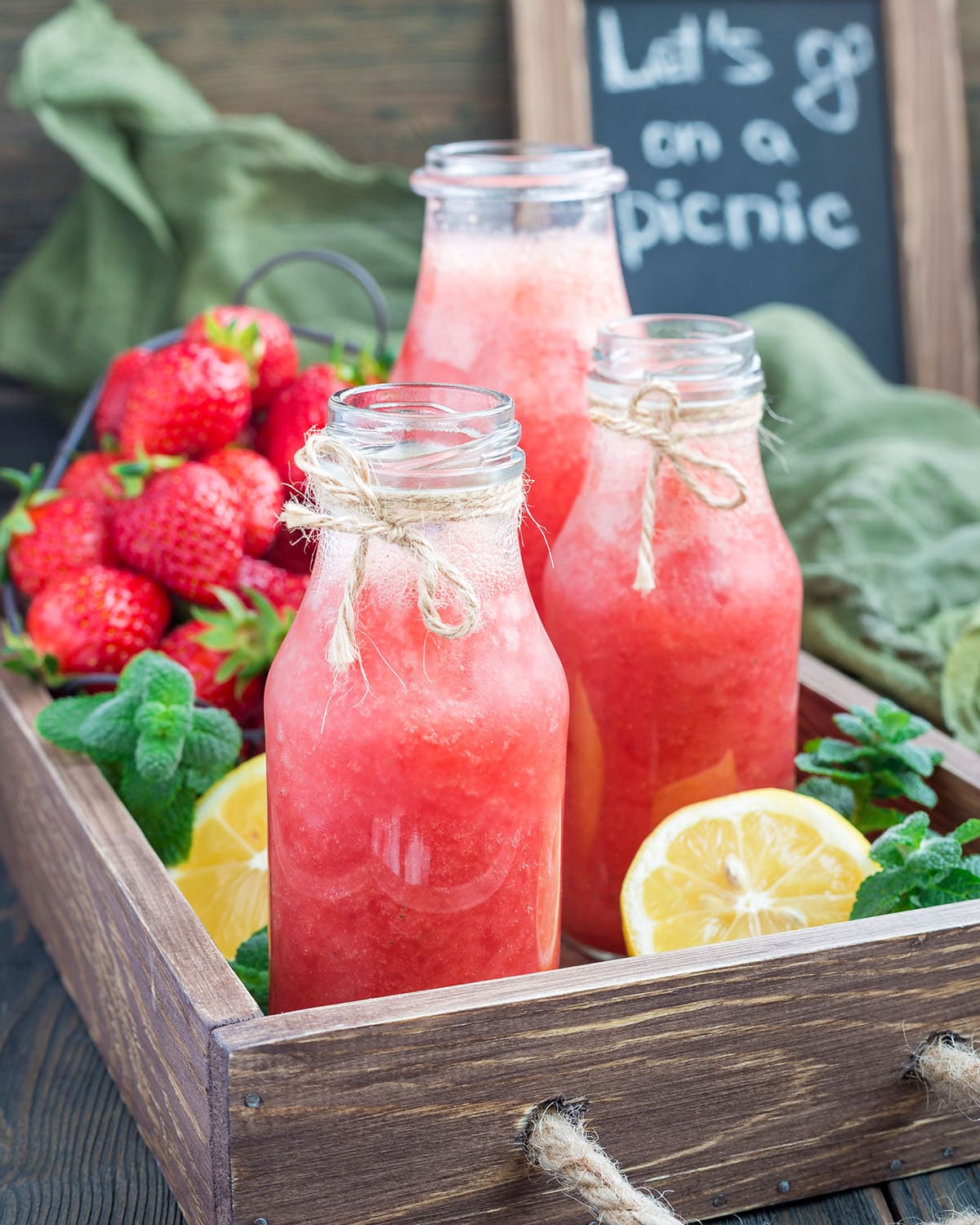 Homemade strawberry lemonade with fresh strawberry, lemon, ice and mint in bottles in a wooden tray with lemons and mint in the background