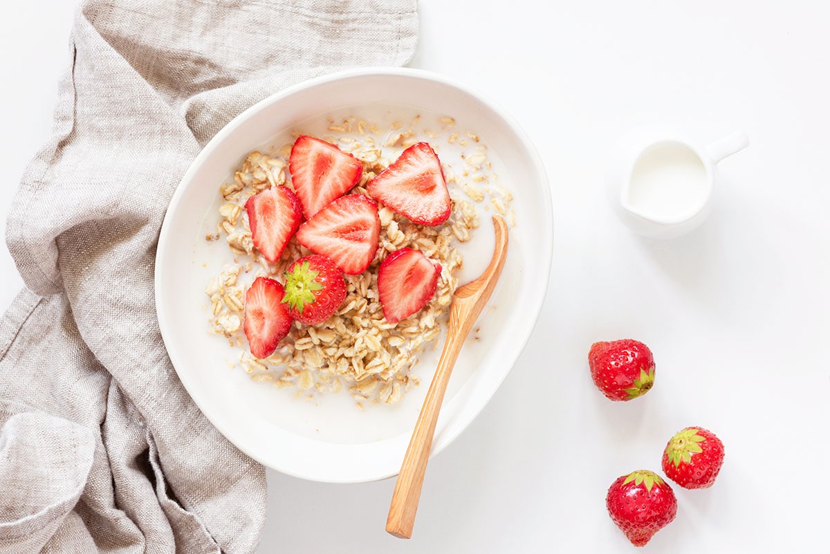 ingredients for strawberry overnight oats: rolled oats, strawberries, milk, yogurt, in a white bowl with a wooden spoon and a gray linen next to the bowl