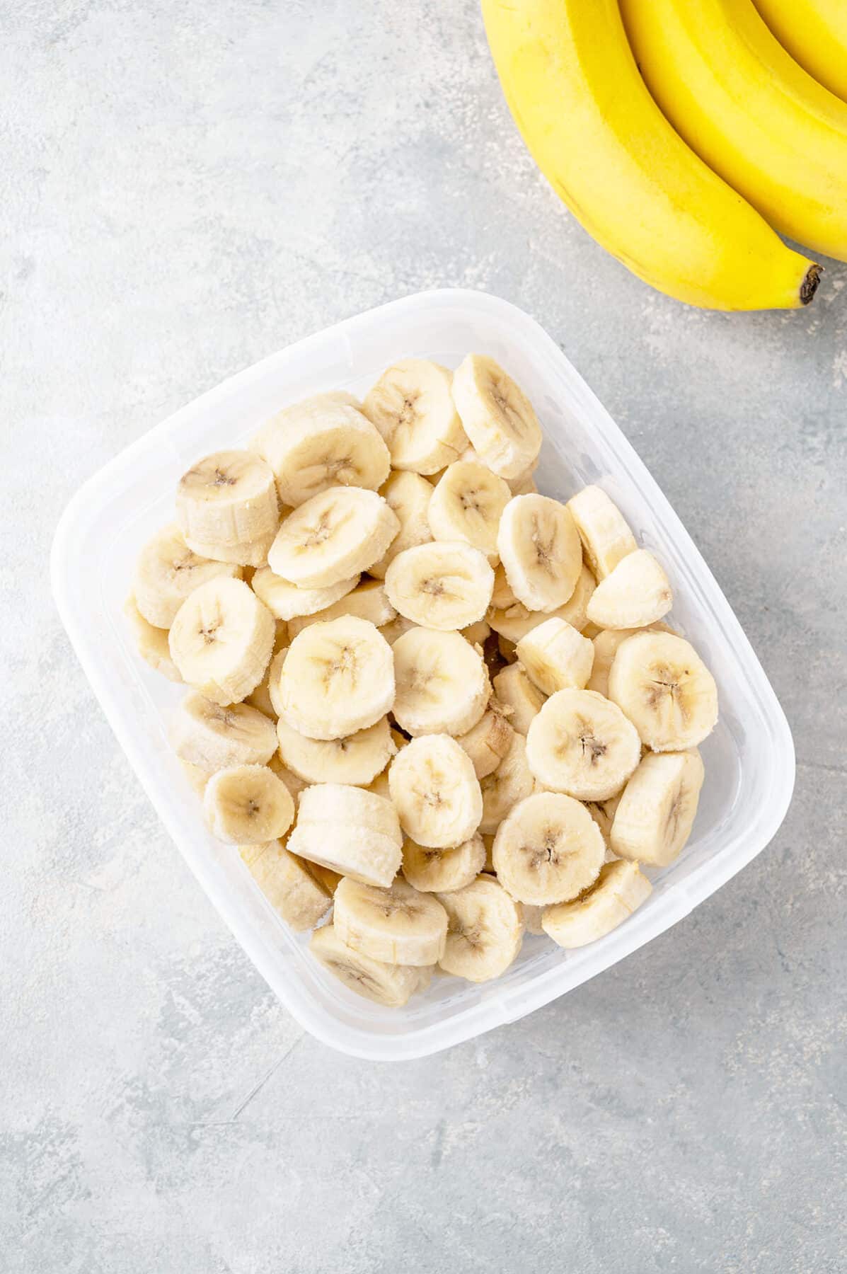 Slices of frozen banana in a container on a gray concrete background.