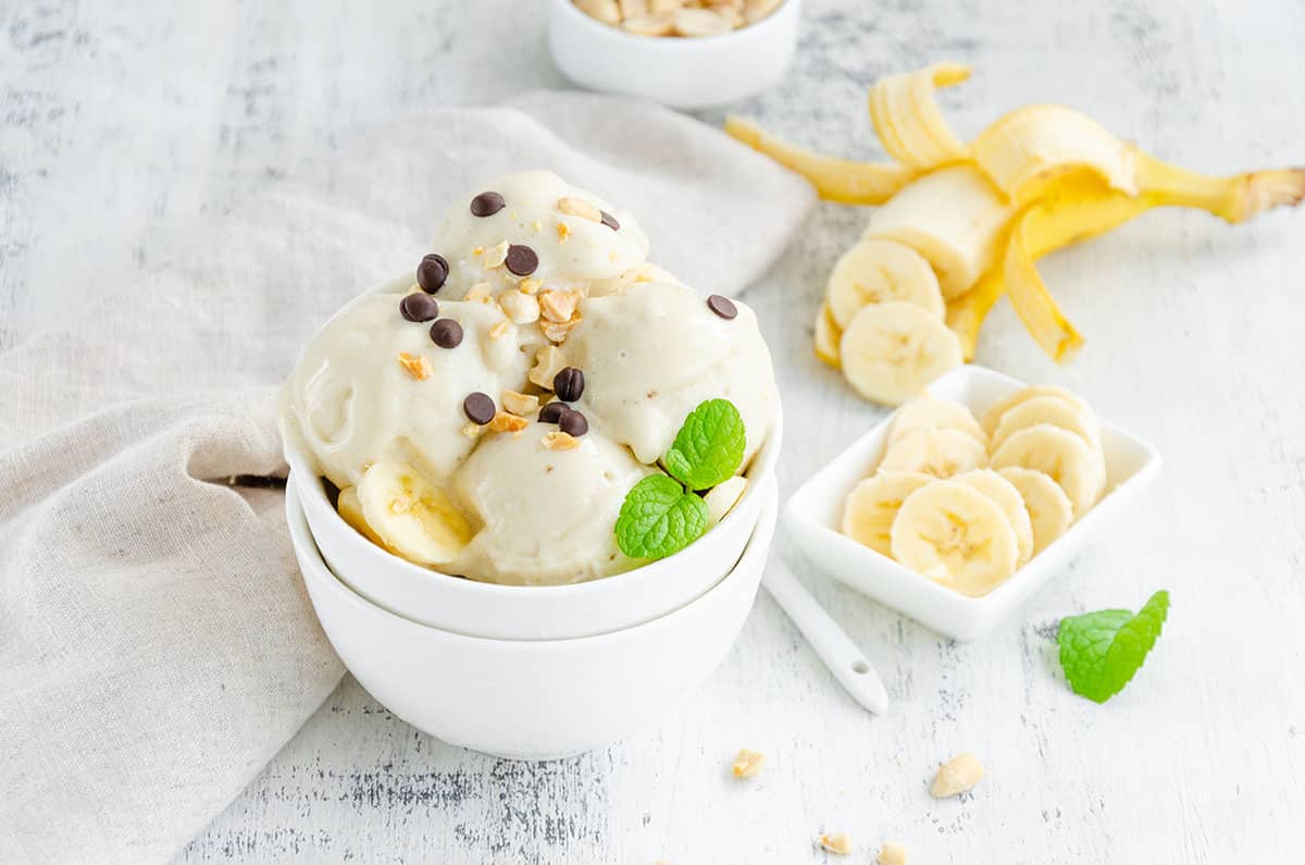  banana ice cream in a bowl with peanuts and chocolate on a wooden background with sliced bananas in the background