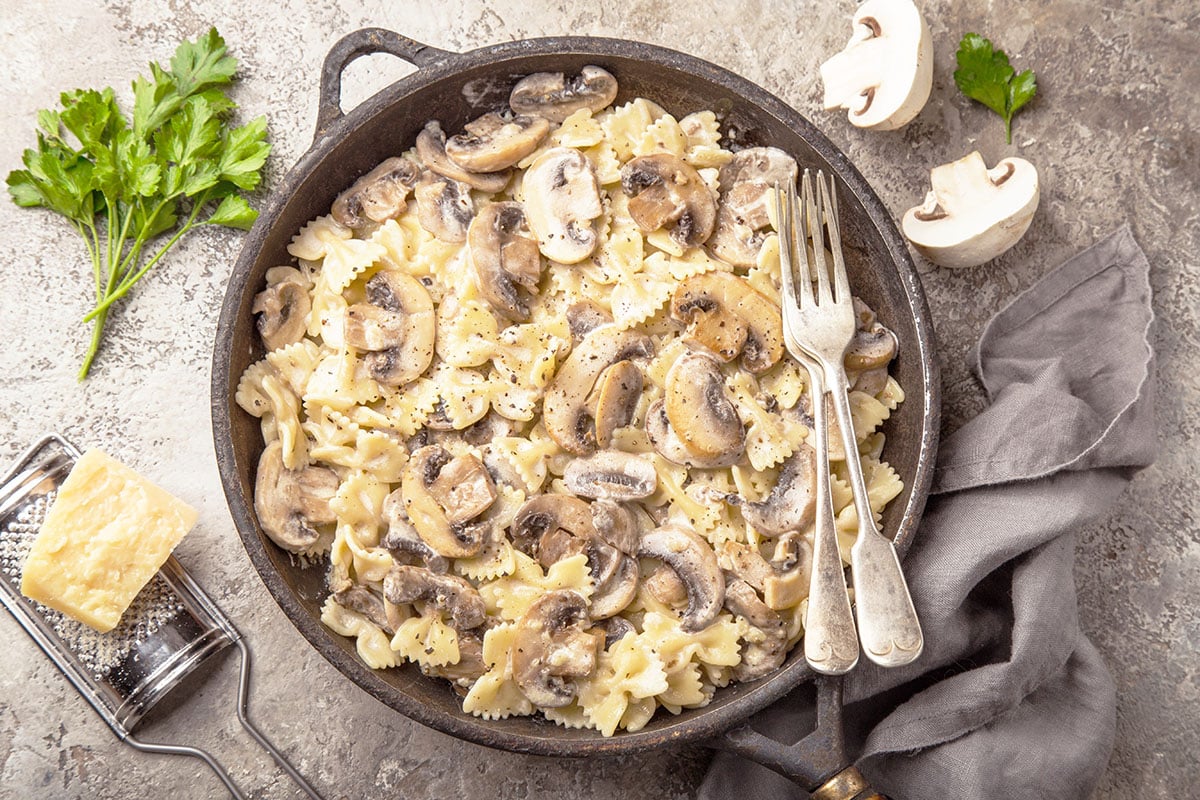 farfalle pasta with mushrooms in a cast iron skillet with parmesan cheese, parsley and mushrooms in the background.