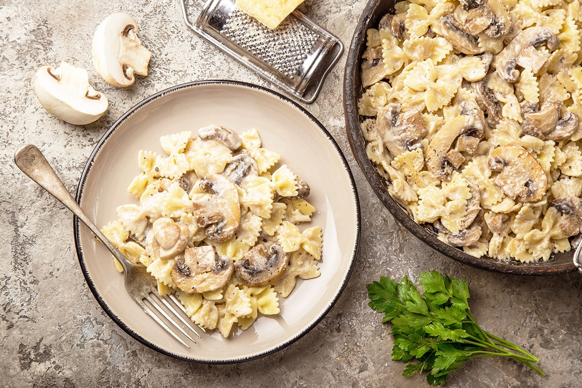 bowtie pasta with mushrooms and a creamy sauce in a cast iron skillet with parmesan cheese, parsley and mushrooms in the background. Plate of mushroom pasta with a fork on the plate.
