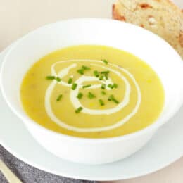 creamy yellow squash soup with a metal spoon and sourdough bread on the sides, green chives on top as a garnish