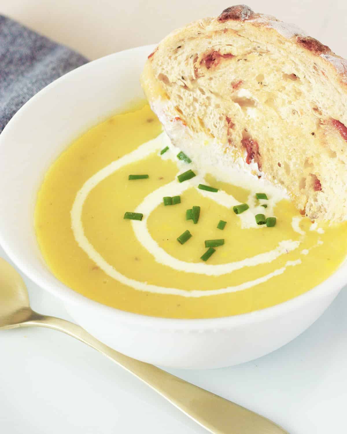 a bowl of yellow squash soup with sourdough bread dunked in the bowl, chives as a garnish and a cream swirl