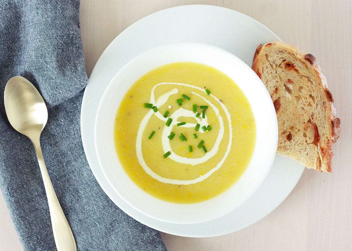 creamy yellow squash soup with a metal spoon and sourdough bread on the sides, green chives on top as a garnish