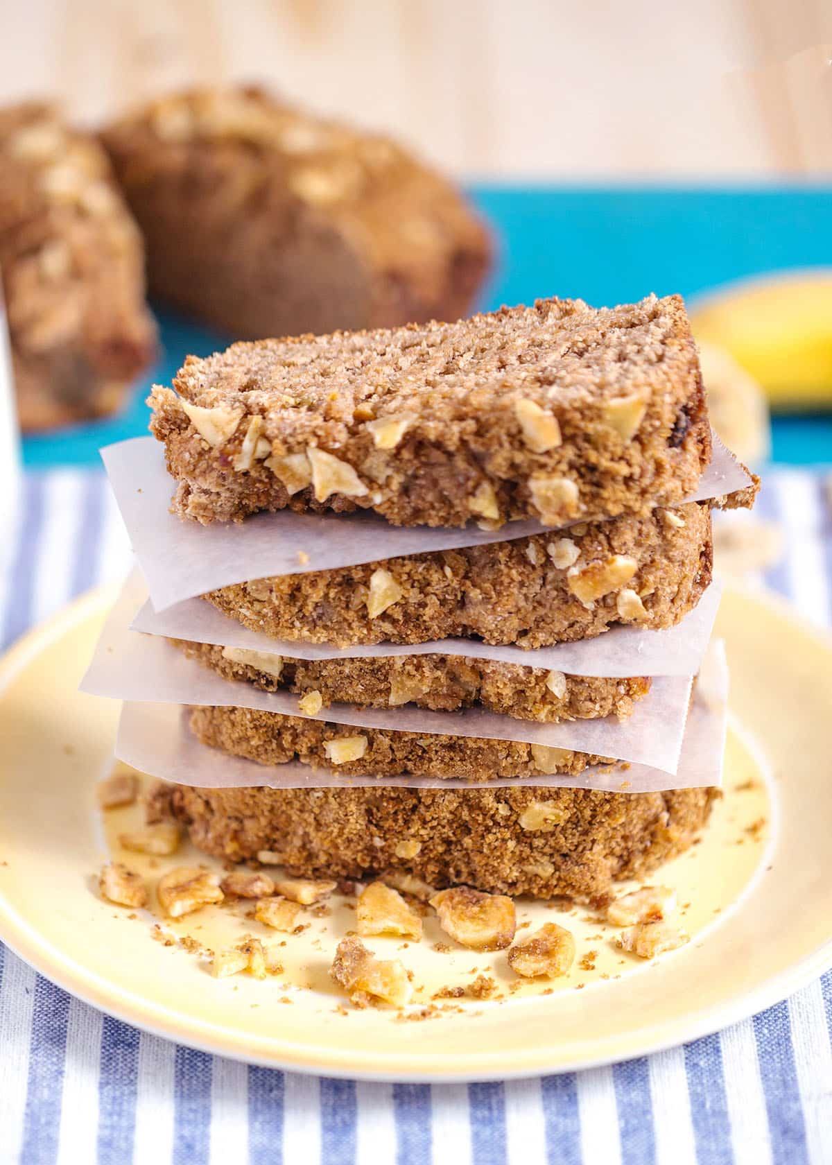 healthy banana bread with streusel topping stacked in slices on a yellow plate with a blue and white striped napkin underneath