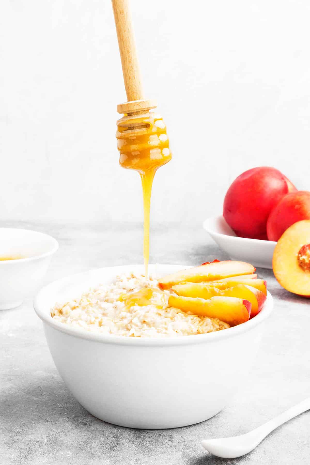 oatmeal with fresh peaches and honey being drizzled into the bowl of oatmeal. Fresh peaches in the background.