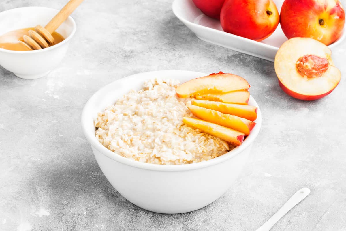 peaches and cream oatmeal with freshly sliced peaches in a white bowl. Honey and whole peaches in the background.