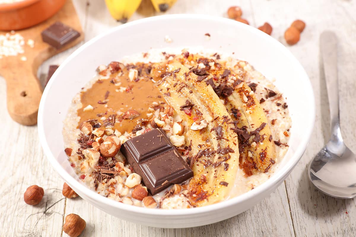 peanut butter banana oatmeal with banana slices, crushed nuts, and shaved chocolate in a white bowl with a wood background