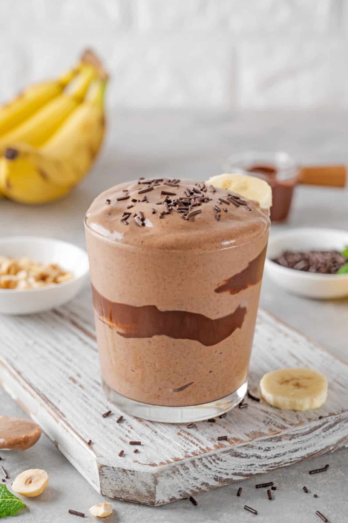 peanut butter banana smoothie with chocolate glaze and sprinkles in a glass on a gray concrete background with bananas and peanut butter.