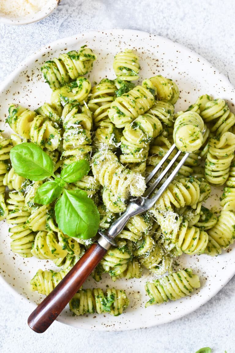 Homemade pesto pasta on a white speckled plate with Parmesan cheese and a fresh basil leaf garnish.