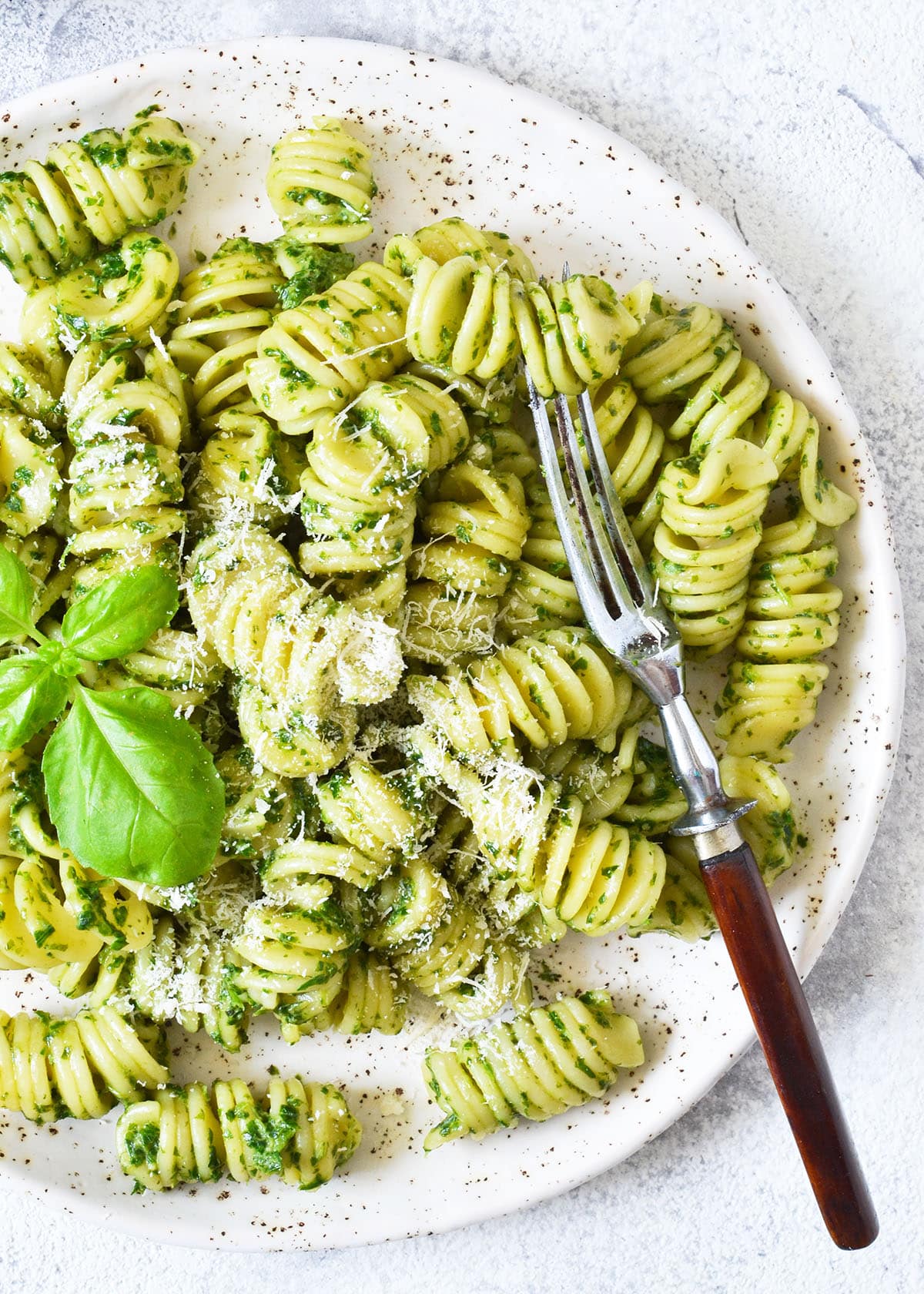 homemade pesto pasta on a white speckled plate with parmesan cheese and basil leaf as garnish.