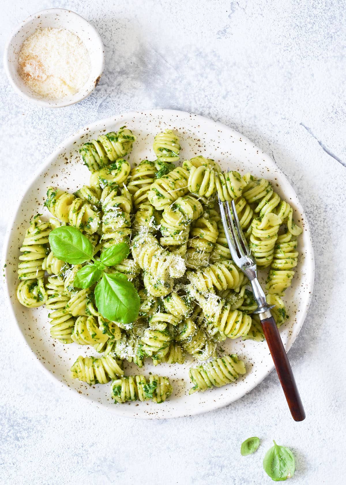 homemade pesto pasta on a white speckled plate with parmesan cheese and basil leaf as garnish.