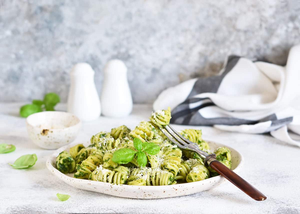 homemade pesto pasta with shredded parmesan cheese and fresh basil as a garnish on a white speckled plate with a fork lifting a piece of pasta. 