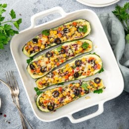 Stuffed zucchini boats with vegetables ( tomato, pepper, corn, red onion and olives) and cheese in white baking dish, top view