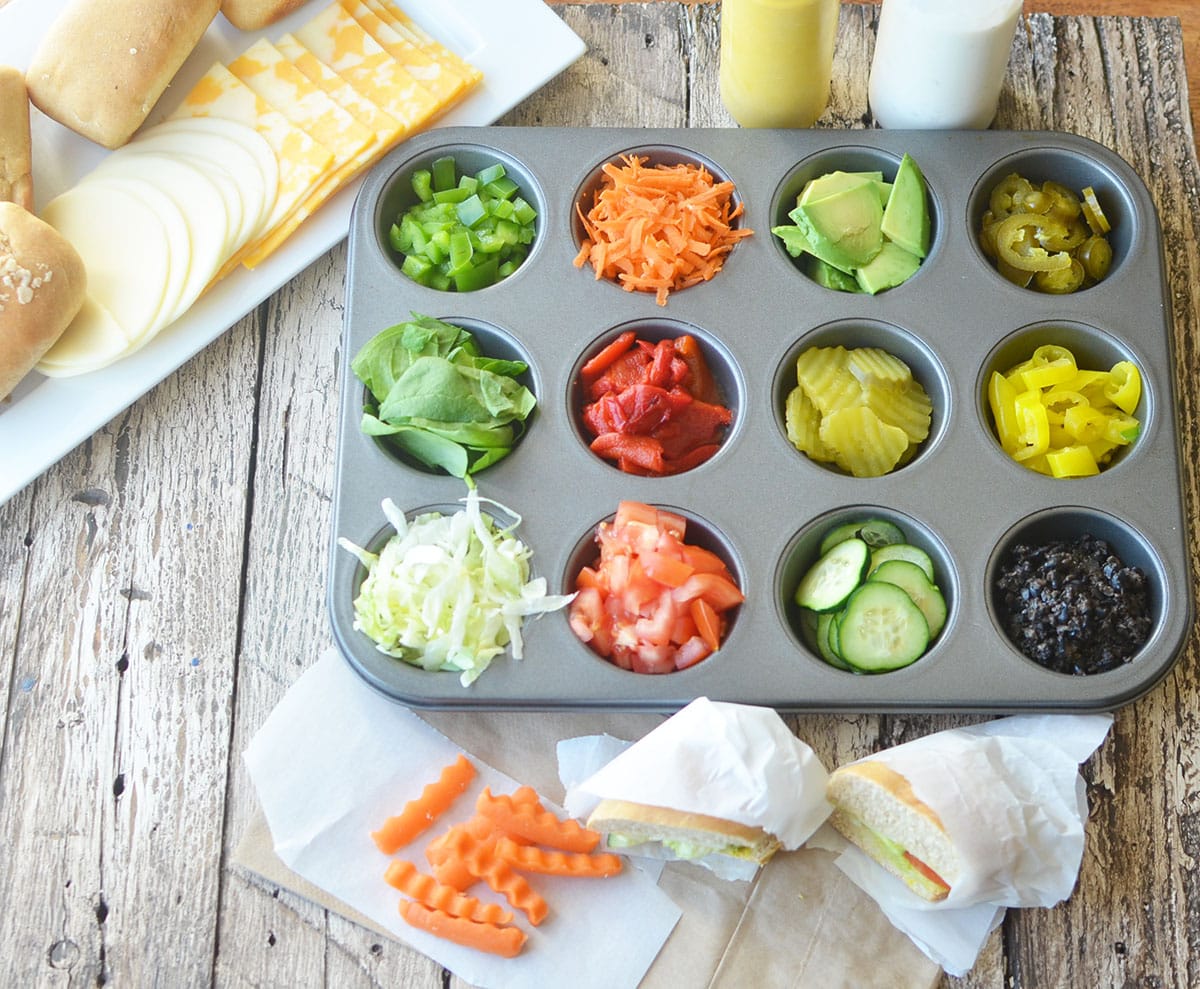 muffin tin with sandwich toppings and cheese and hoagie rolls in the background