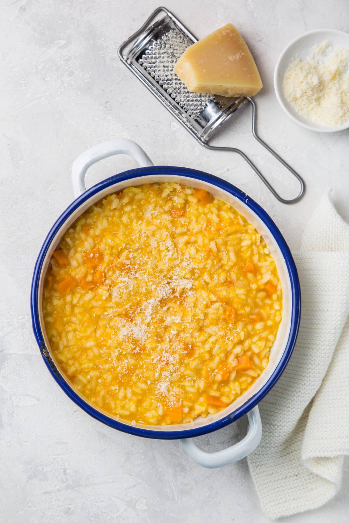 Butternut squash risotto in a white and blue ceramic pot with cheese and a grater in the background.