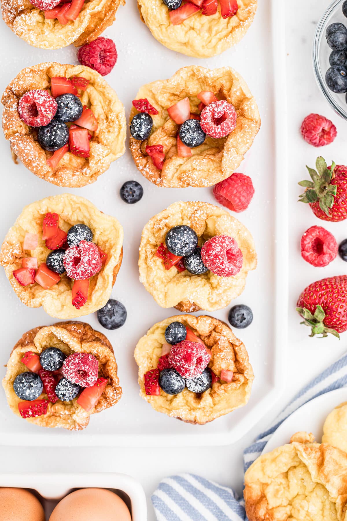 overhead view of popovers on a white tray with berries and powdered sugar on top and berries sprinkled on the counter