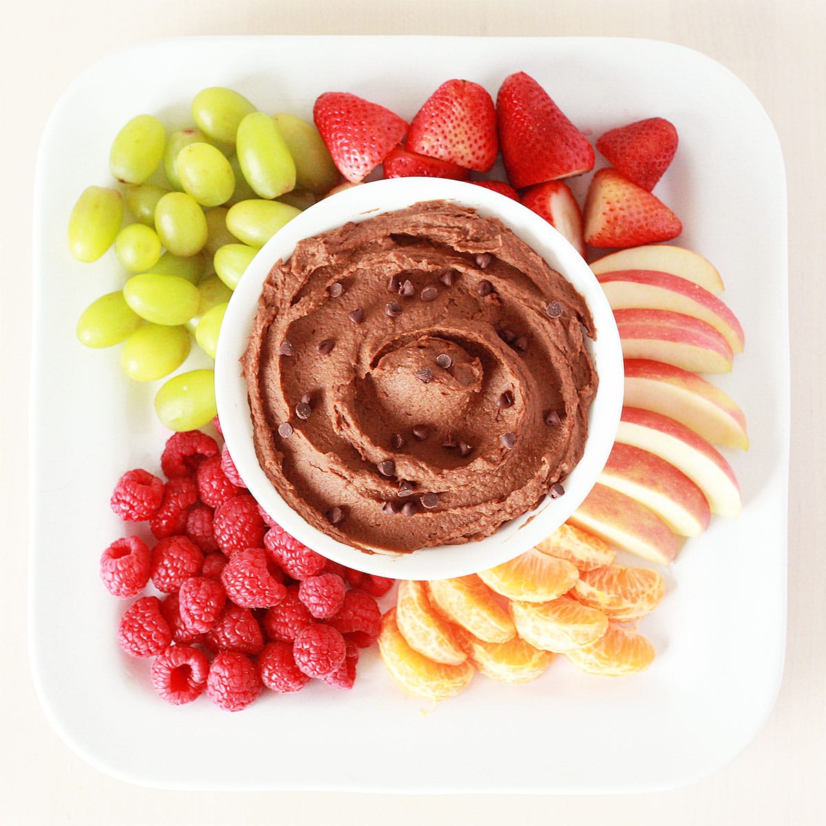 a bowl of chocolate hummus topped with chocolate chips on a white platter with fresh fruit