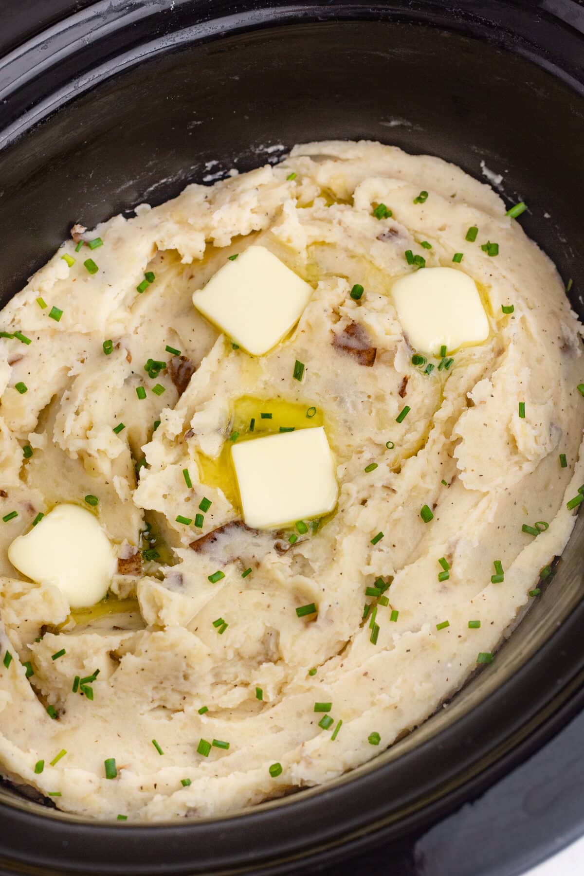 mashed potatoes in the crockpot with chives and butter on top in a black crockpot