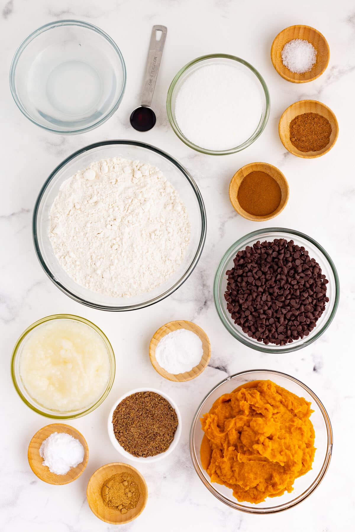 All of the ingredients for pumpkin muffins in individual bowls on a white counter