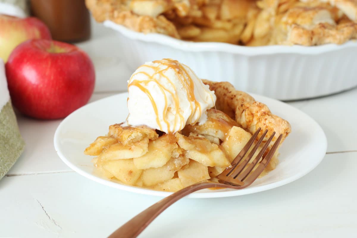 a slice of caramel apple pie with vanilla icecream on top and an apple and the pie in the background