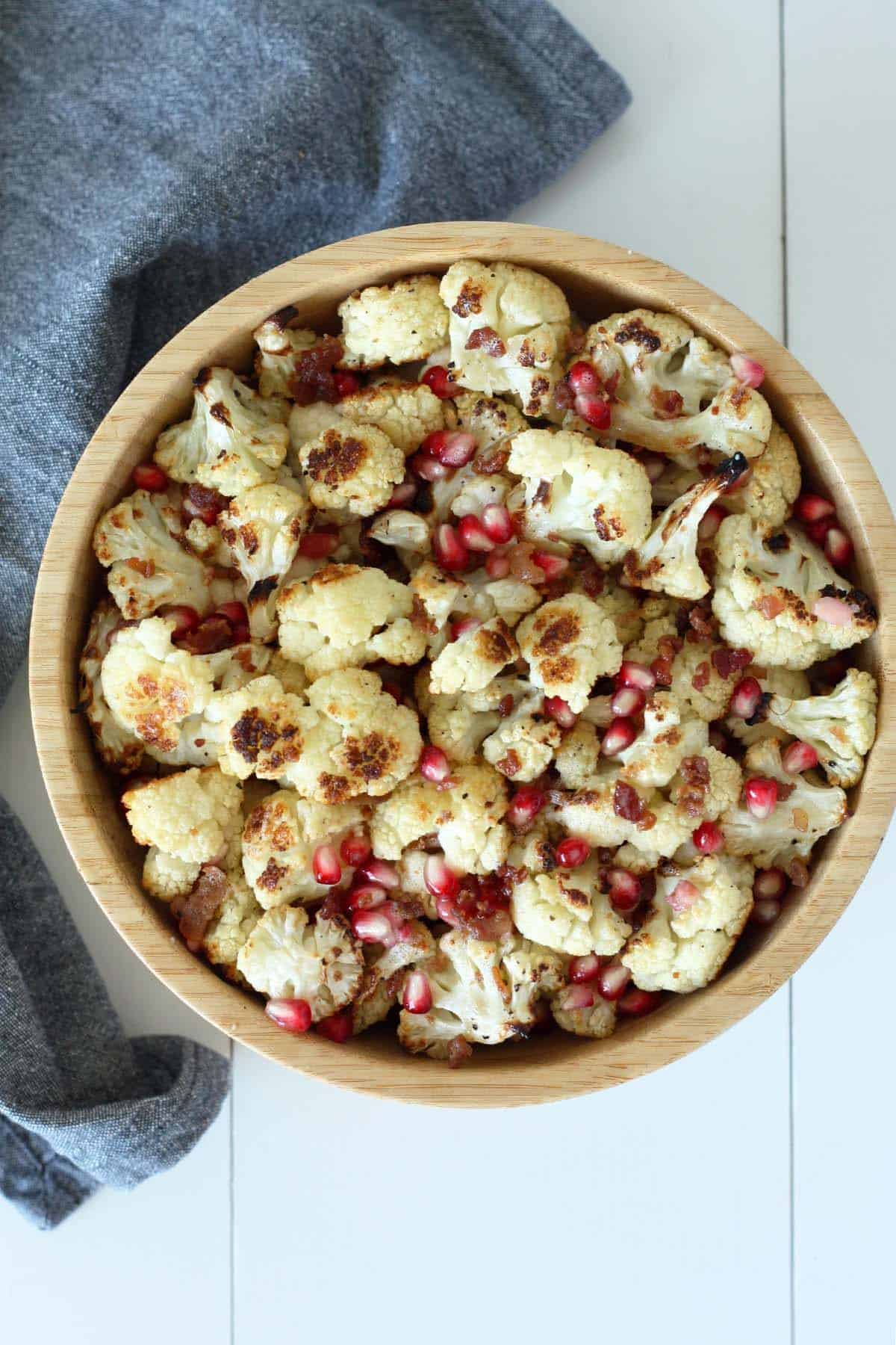 overhead view of roasted cauliflower in a wood bowl with bacon pieces and pomegranate arils and a blue linen in the background