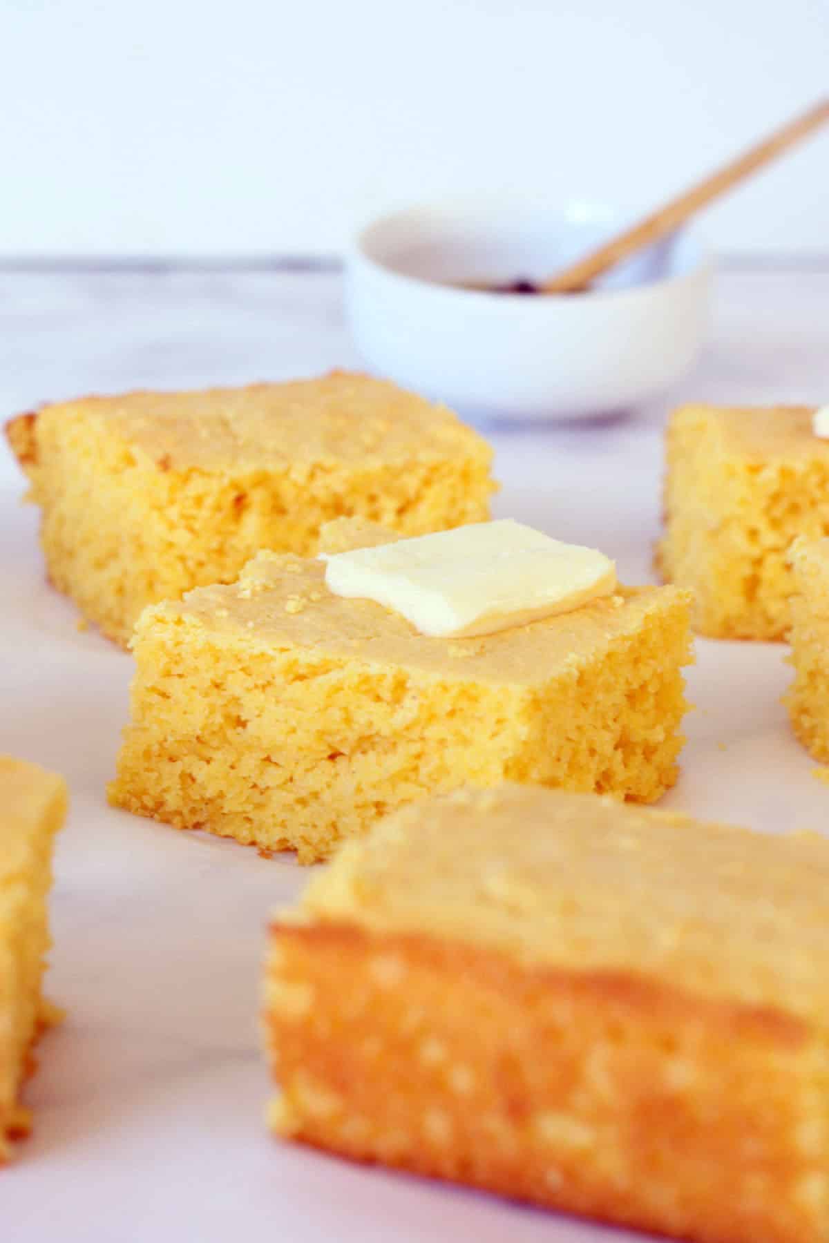 golden sweet cornbread squares on parchment with a pat of butter on top and honey bowl in the background