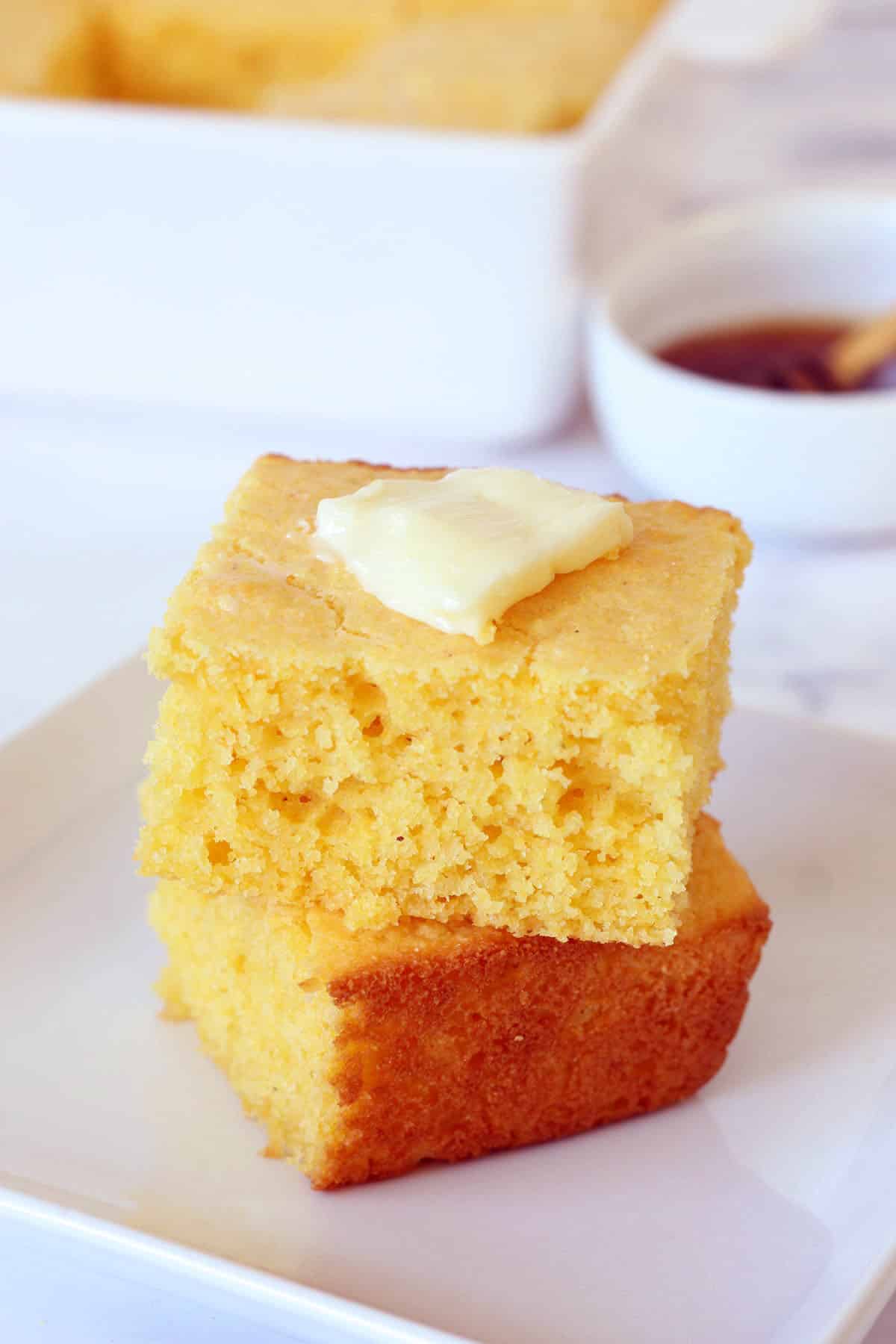 cornbread squares stacked on a white plate with a pat of butter on top and a bowl of honey in the background
