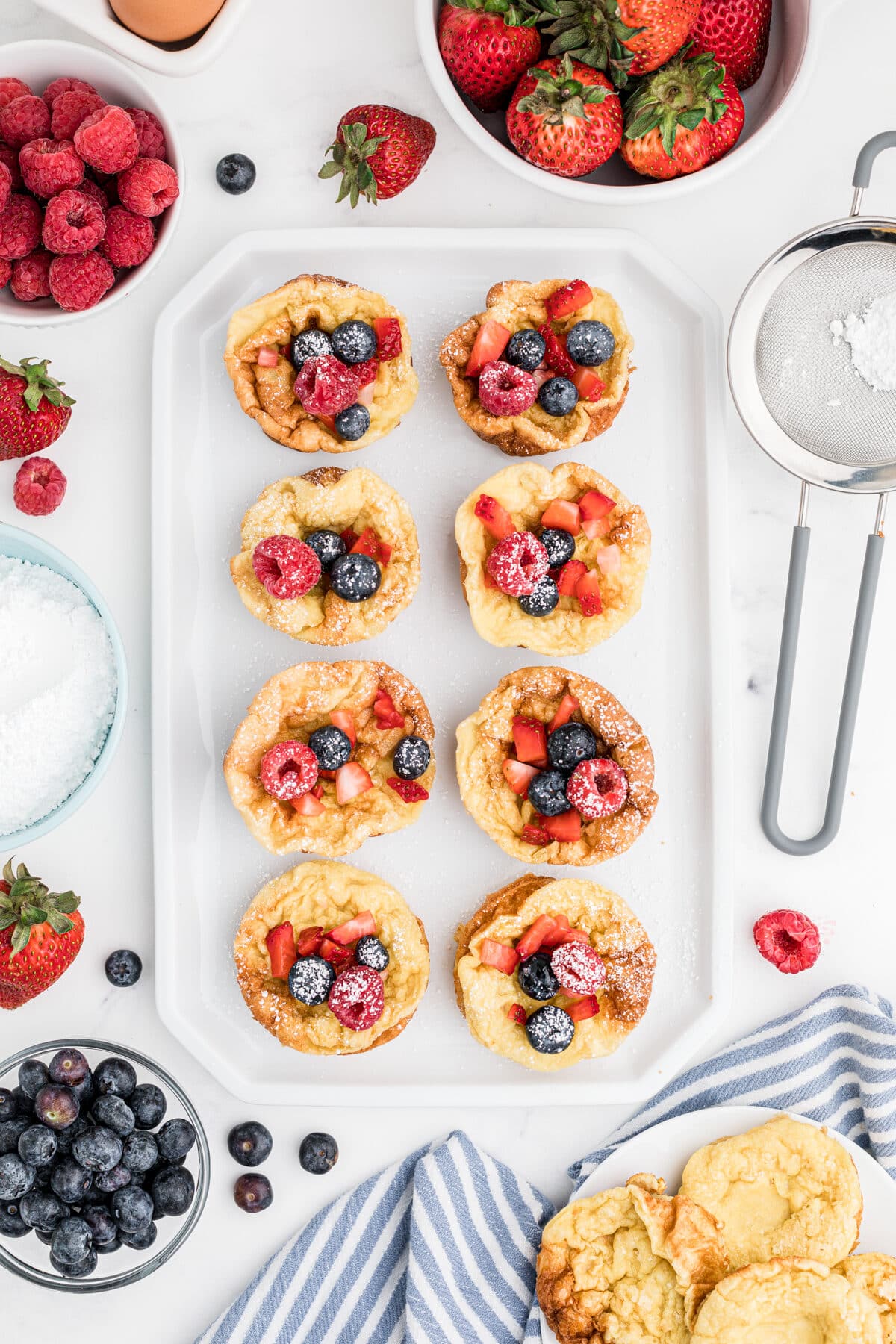 overhead view of popovers on a white tray with berries and powdered sugar on top and berries sprinkled on the counter