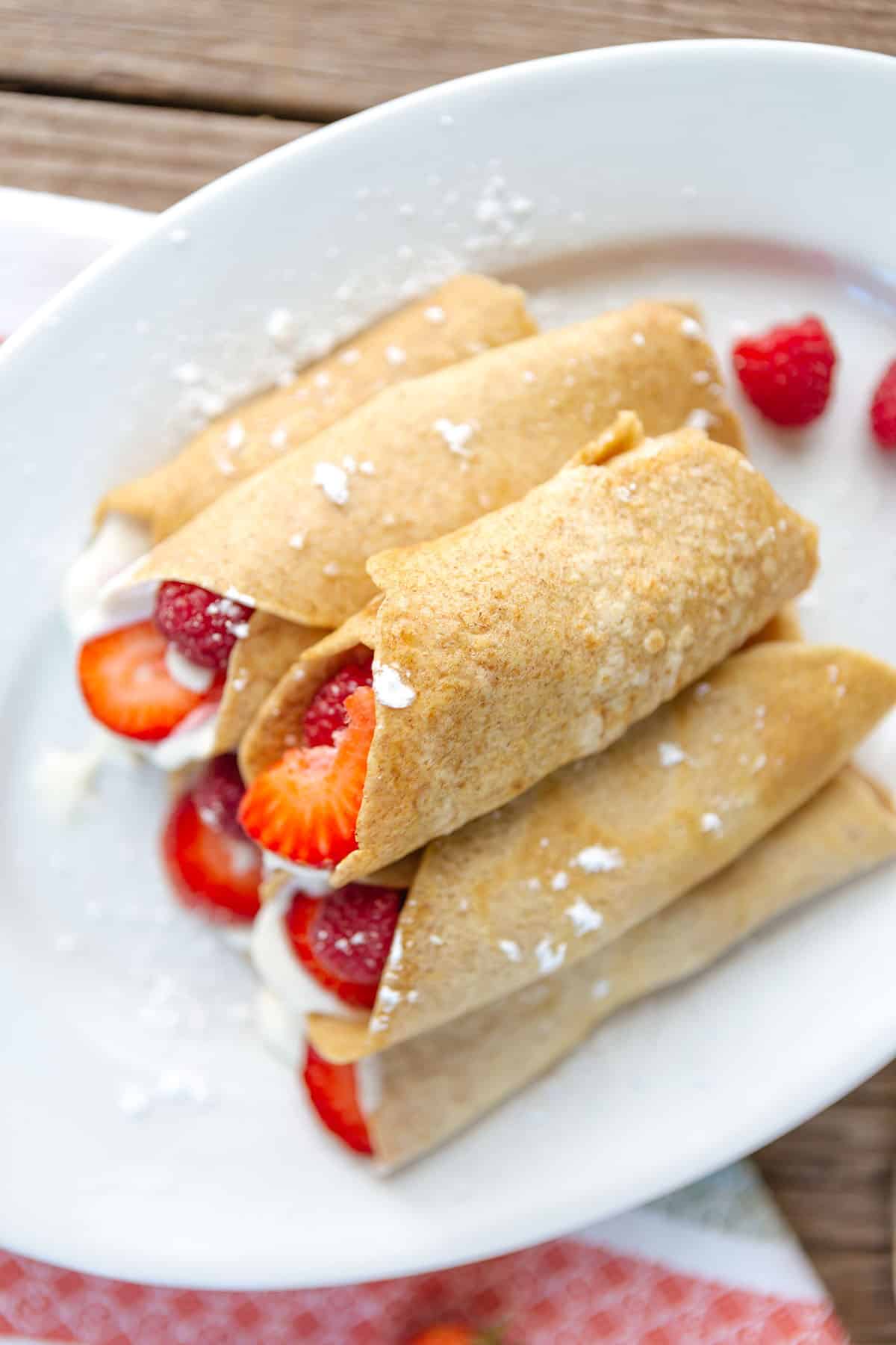 An overhead view of strawberry crepes filled with fresh strawberries and raspberries and cream filling on a white plate