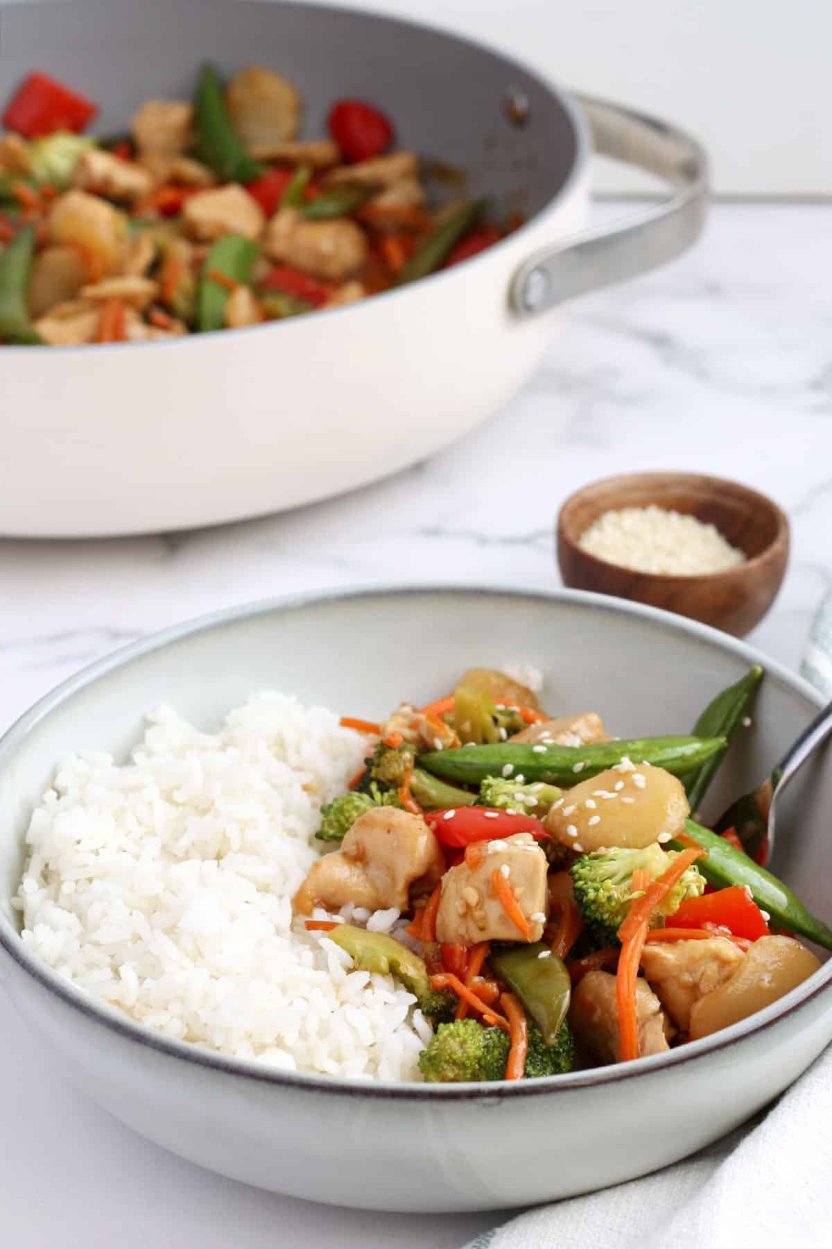 a large serving bowl with rice and stir fry chicken next to a small wooden bowl of sesame seeds