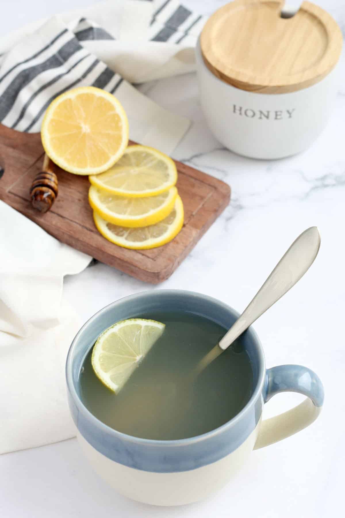 Blue mug and silver spoon with honey lemon cold medicine next to cutting board with lemon slices