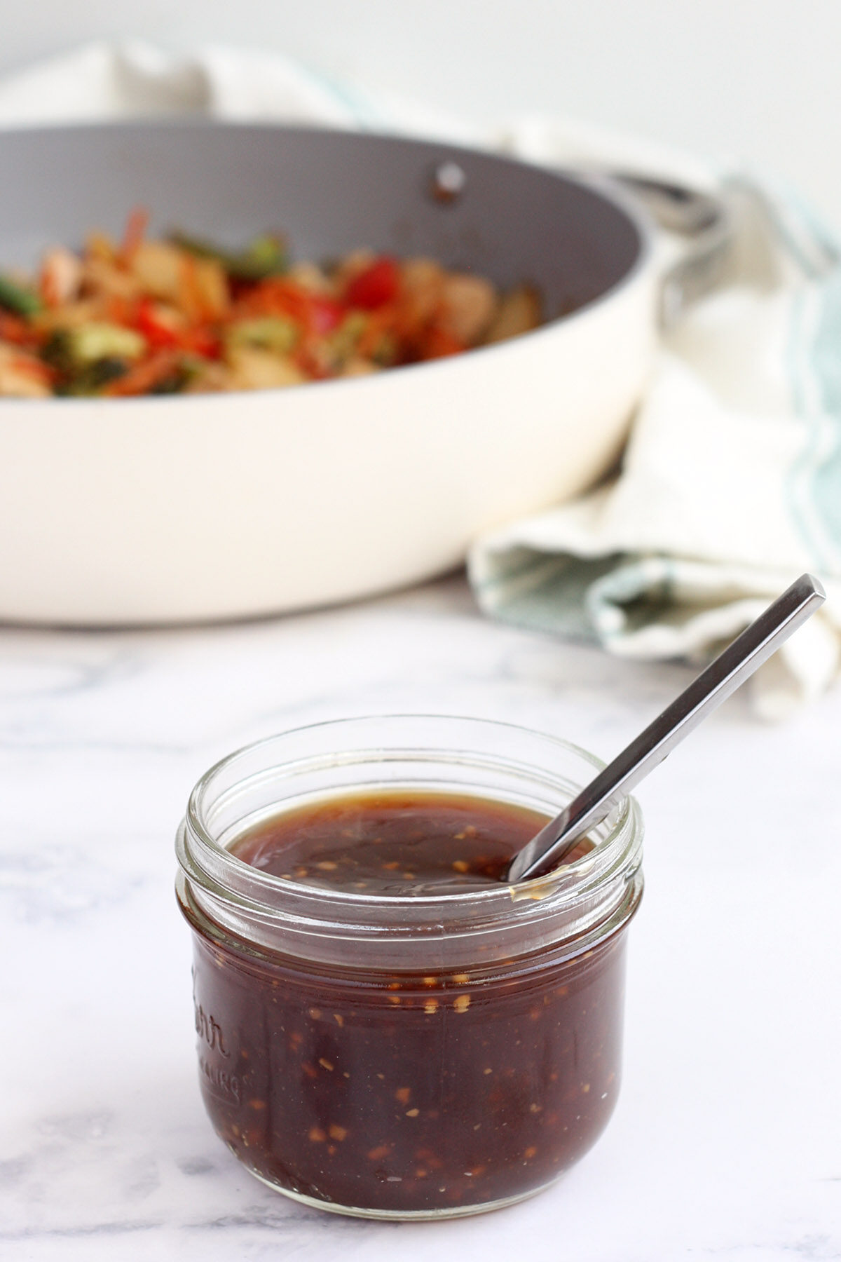 a jar of homemade stir fry sauce with a spoon in front of a stir fry pan