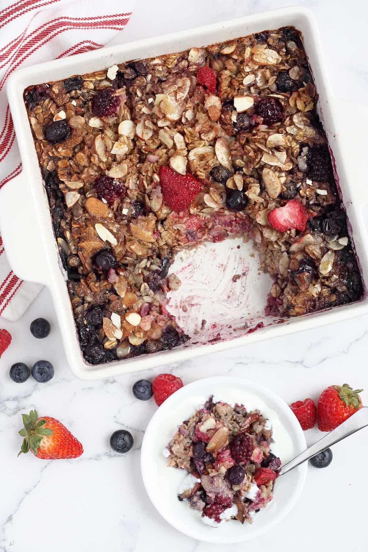 Overhead shot of a baked berry oatmeal casserole with a serving on a white plate.
