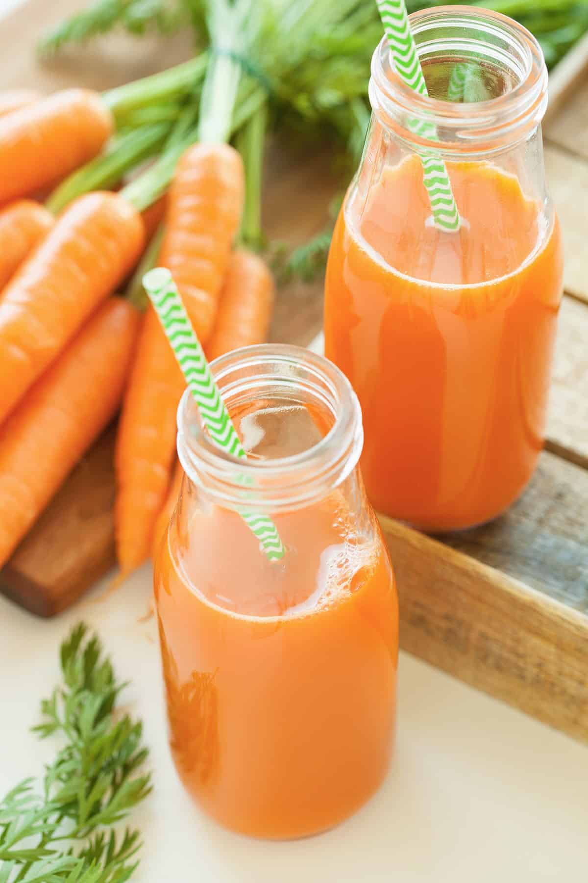 Carrot juice in glass bottles with green striped straws and fresh carrots on background