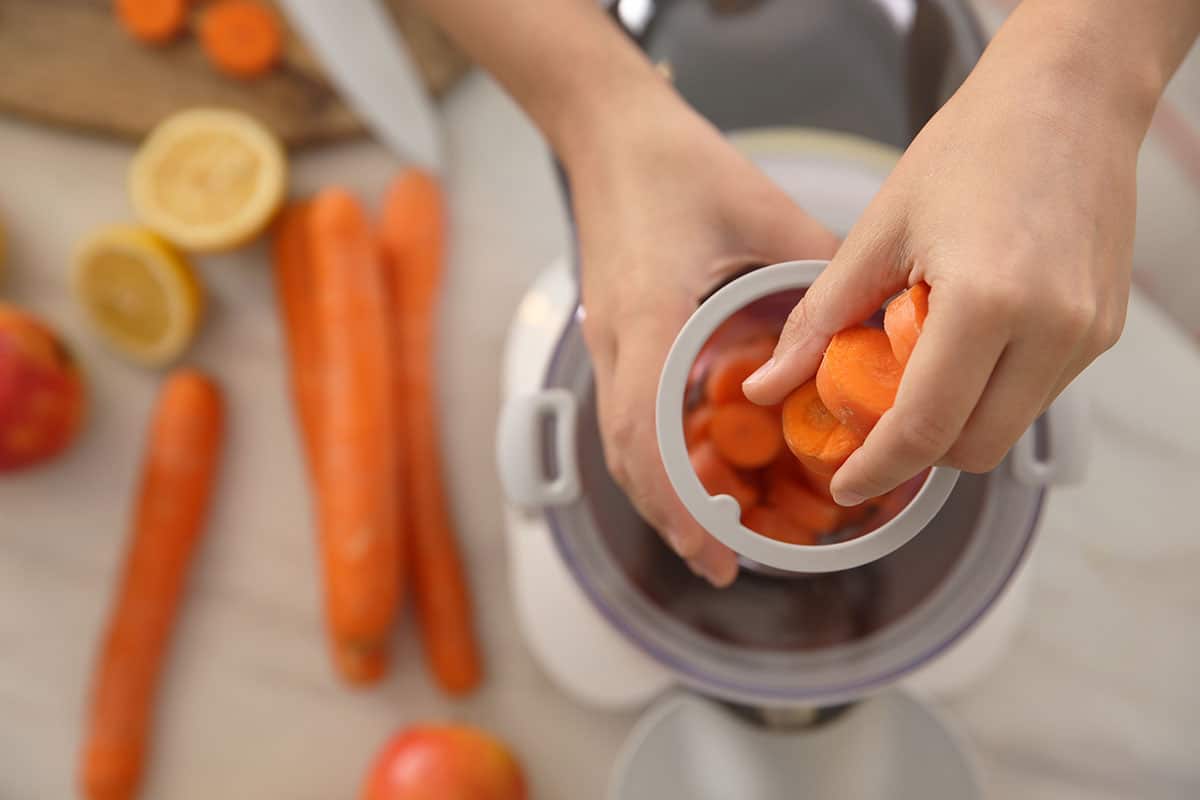 Young woman putting fresh slices of carrot into juicer at table, top view