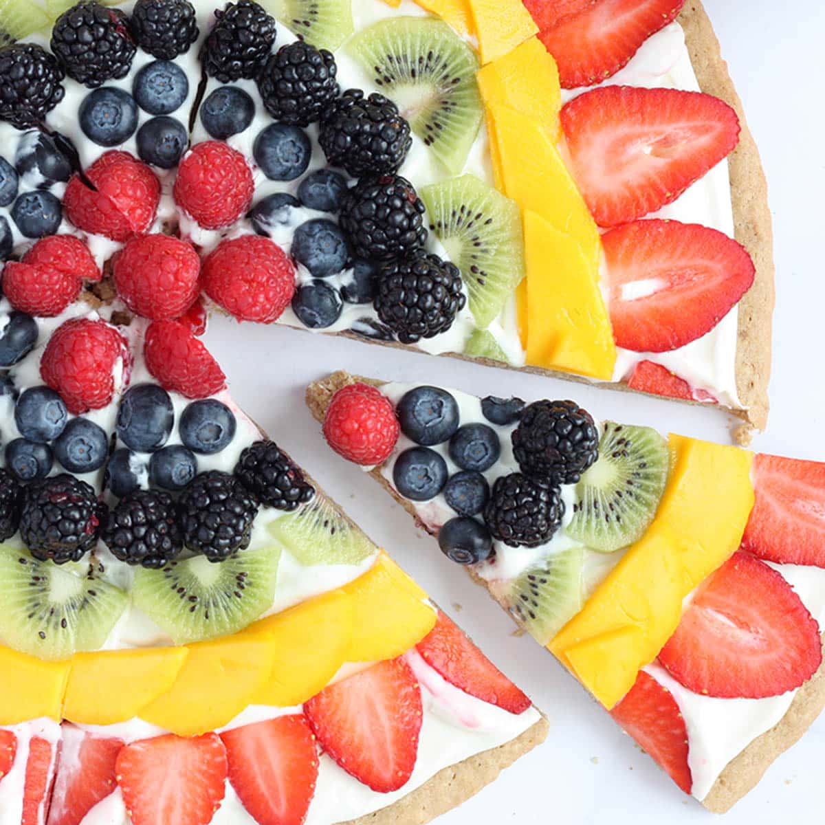 fruit pizza with a slice cut out with strawberries, mango, kiwi, blackberries, blueberries, raspberriiess
