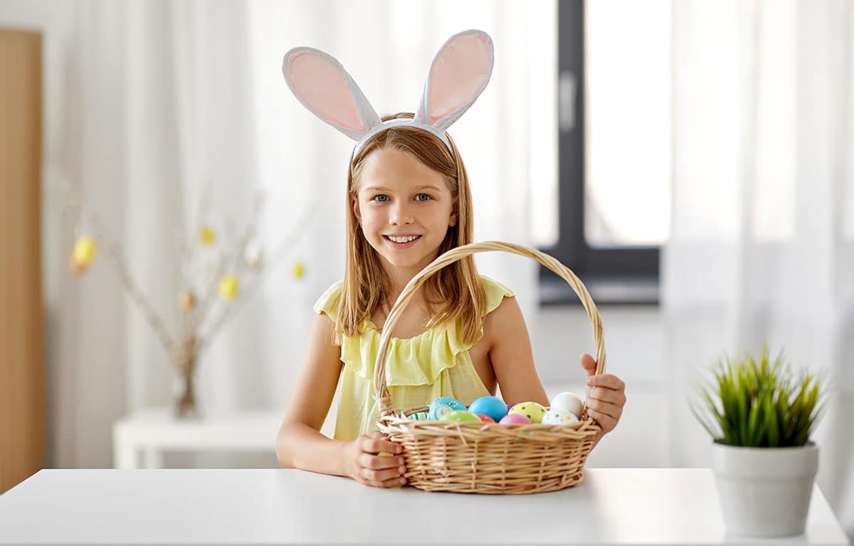 easter, holidays and people concept - happy girl wearing bunny ears headband with basket of colored eggs at home