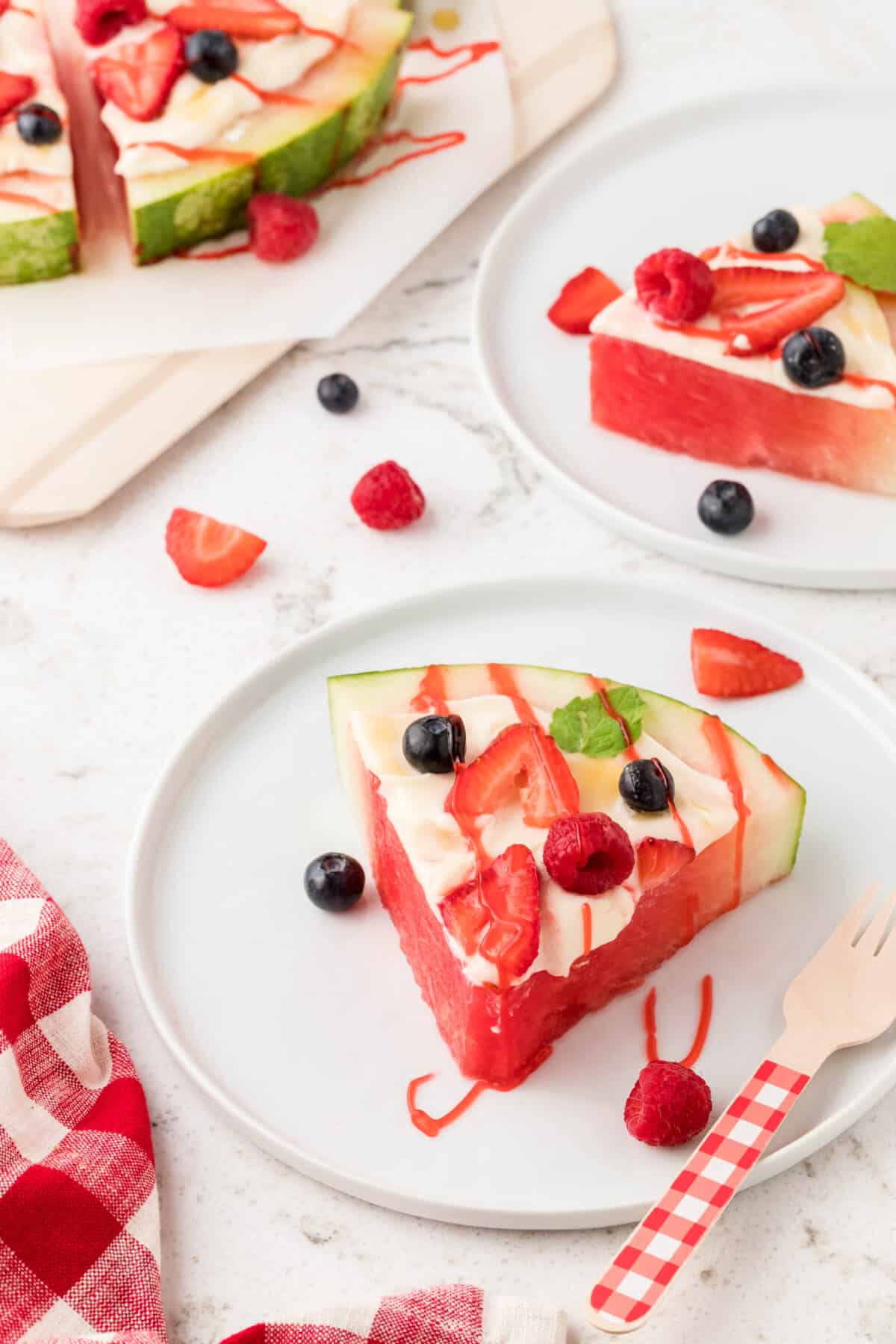 A slice of watermelon topped with whipped cream, fresh berries and a strawberry drizzle on a serving plate.