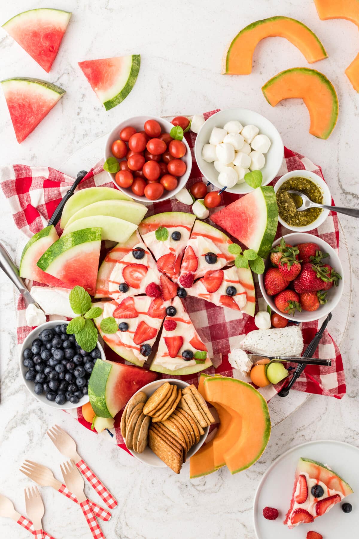 A summer charcuterie board with watermelon pizza, fresh fruit, cheese and crackers on a red checkered linen.