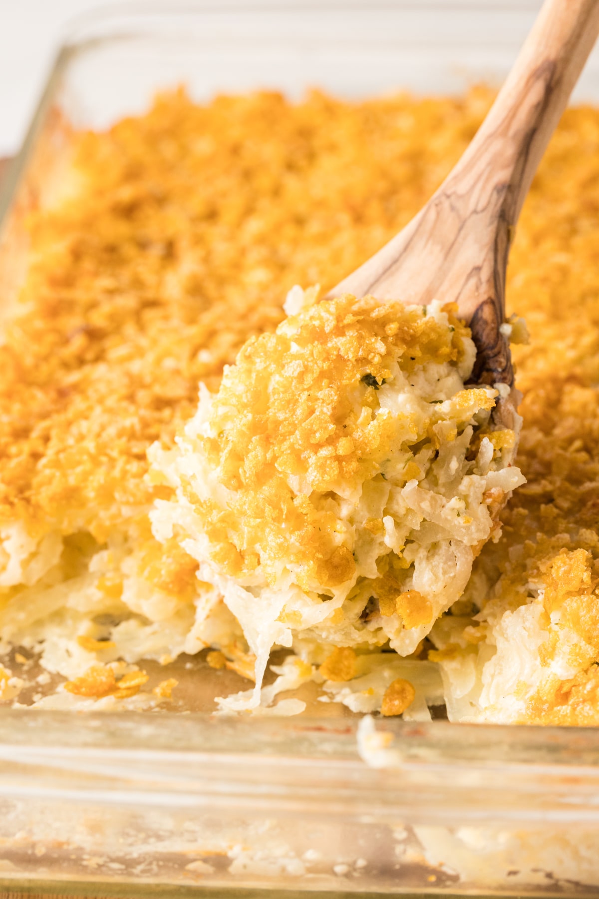 cheesy potatoes being scooped out of a glass casserole dish with a wood spoon